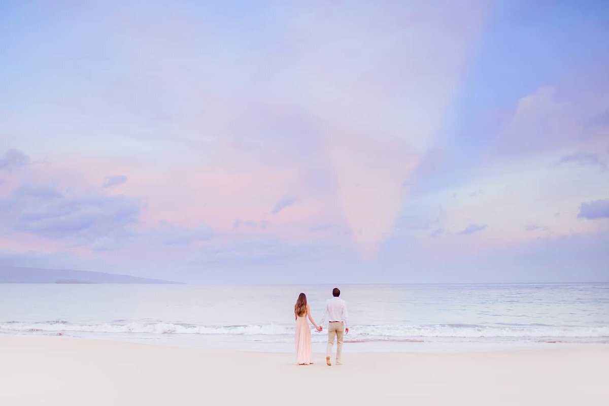 Young couple walks away from the camera toward the ocean on Maui during their sunrise beach engagement portraits in south Maui