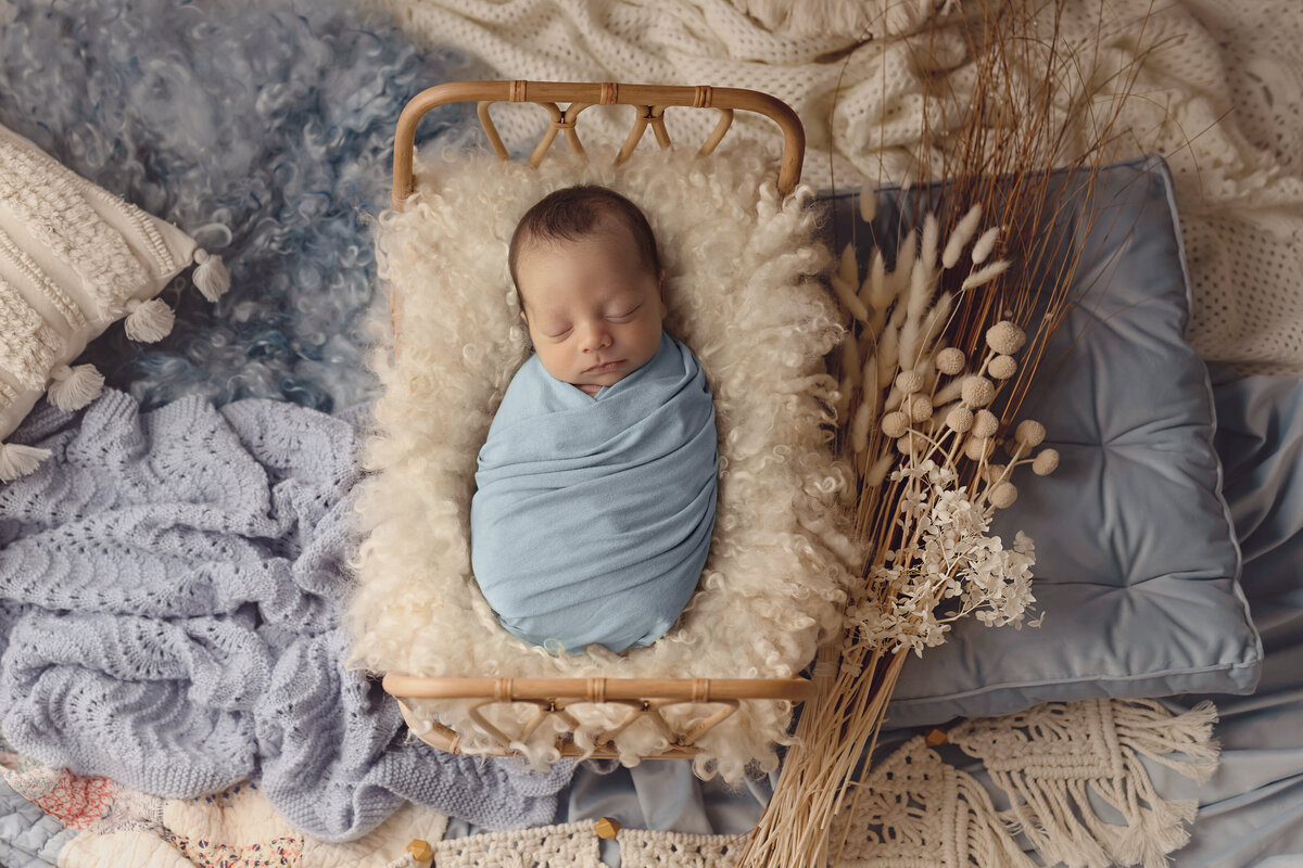 A newborn baby sleeps in a wicker bed on the floor of a studio covered in blankets and pillows