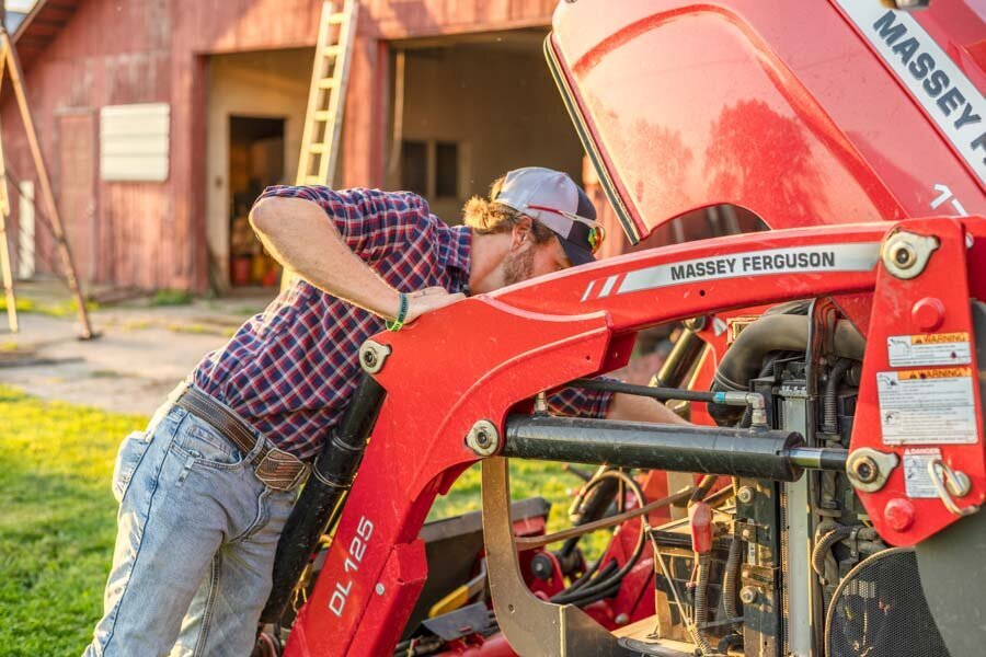 A man in a plaid shirt and jeans inspects the engine of a red massey ferguson tractor near a barn.
