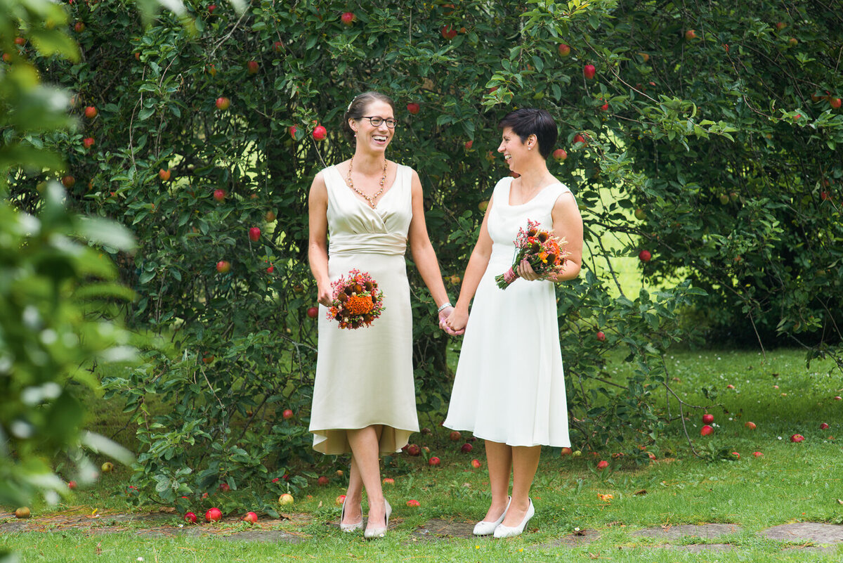 Gay wedding couple wearing white dresses, standing in garden , holding hands and colourful bouquets