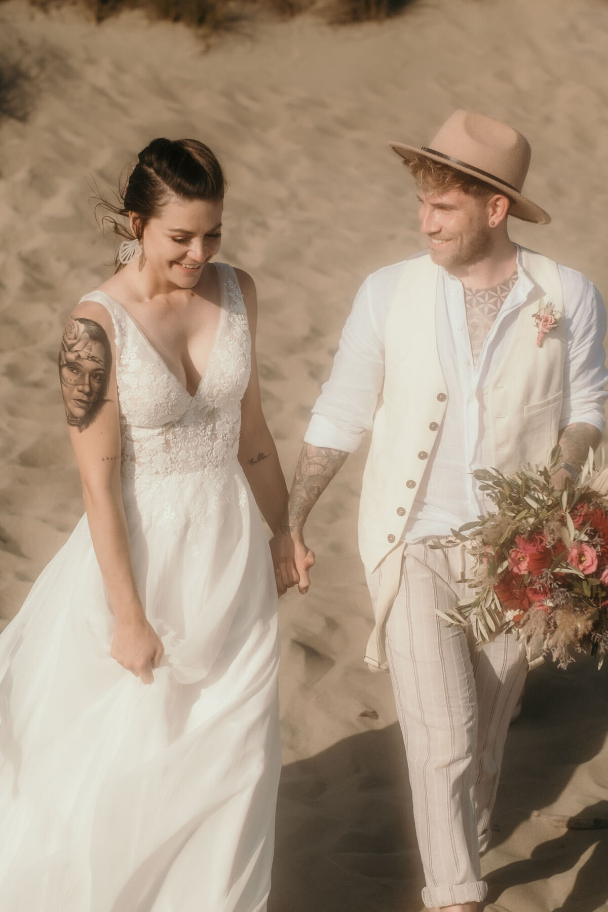 Glücklich lächelnd läuft das Hochzeitspaar Händchen haltend im Sand. Der Bräutigam sieht die Braut an.