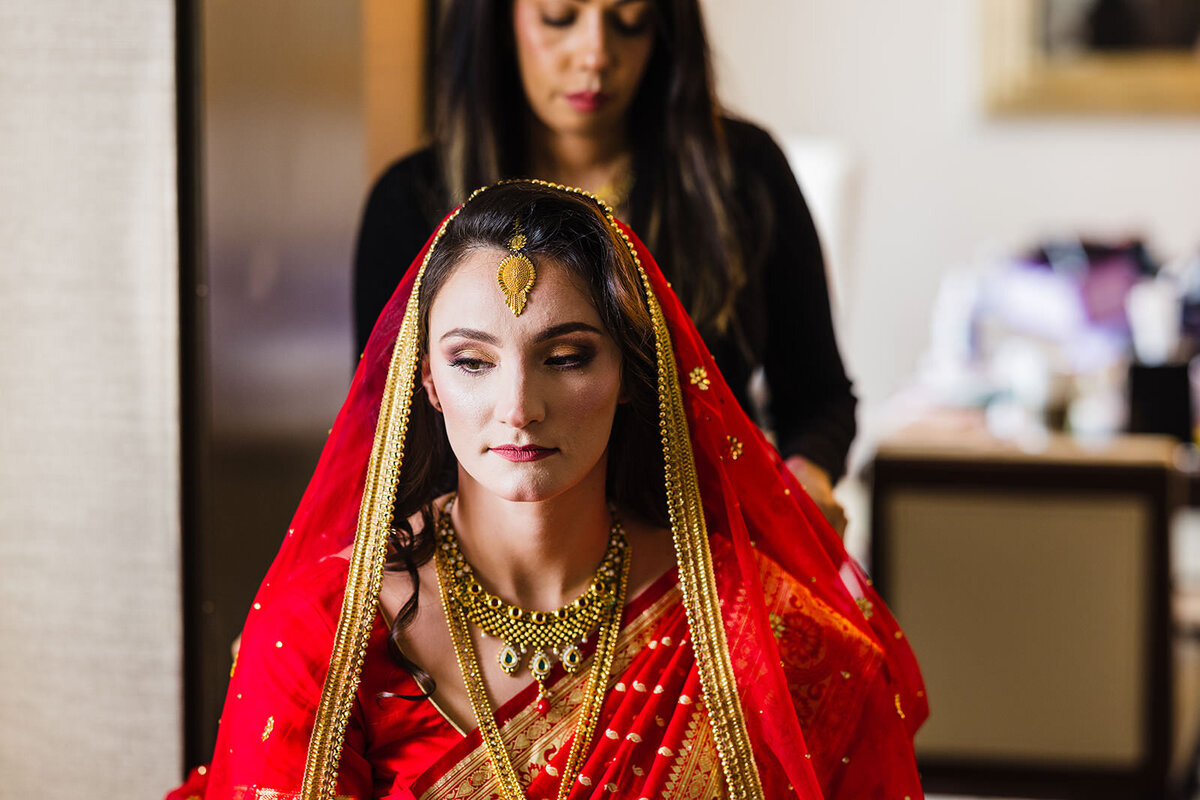 A close-up of a bride in a red bridal attire with elaborate golden jewelry, her face framed by the sheer red dupatta.