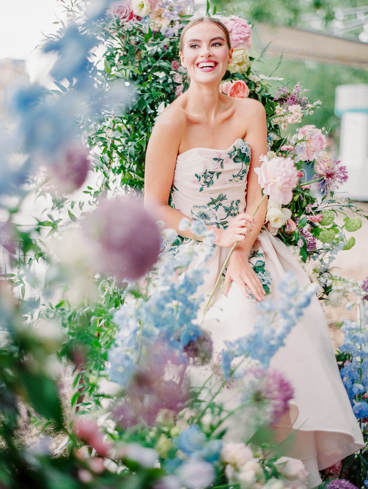 Bride at pre-wedding bridal session in Paris on the Seine River surrounded by colorful florals while wearing a strapless pink gown with embroidered colorful flowers photographed by Paris wedding photographer