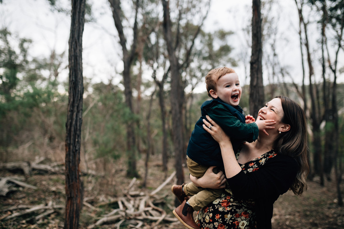 woman holding young son up in the air, laughing together, outside in bushland.