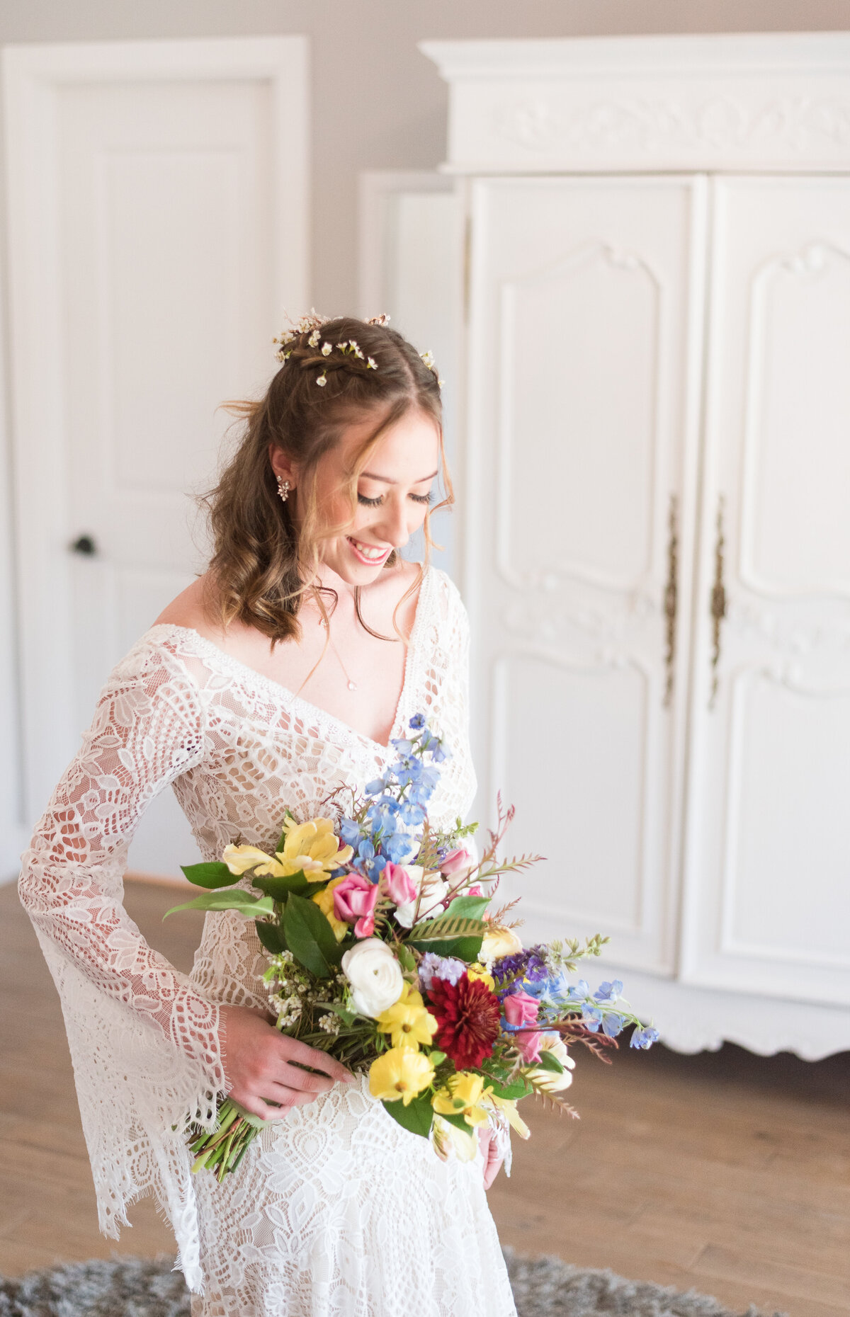 bridal portrait of bride in a lace boho wedding dress in her bridal suite holding her bright and colorful floral bouquet as she smiles while looking down at her wedding flowers