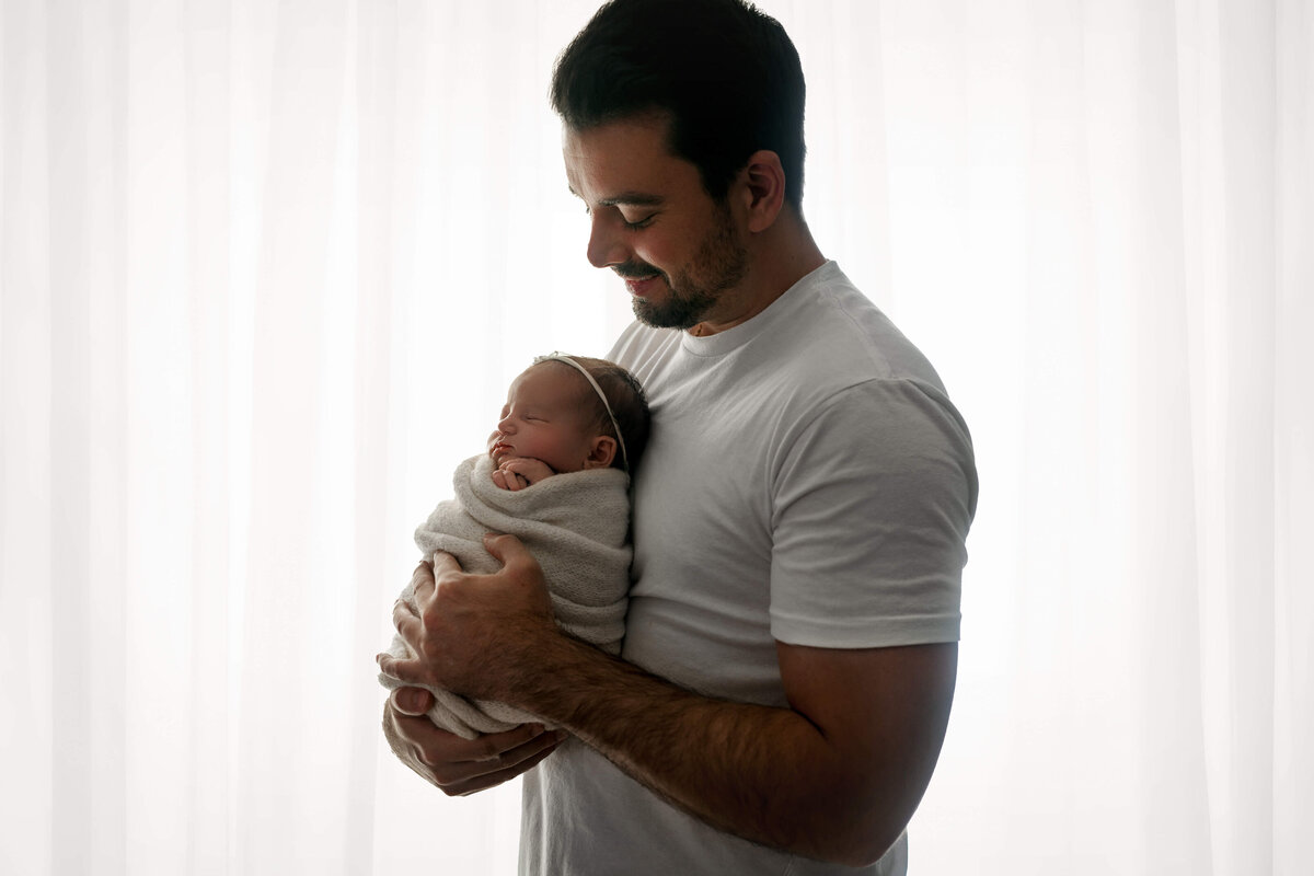 new dad holding his newborn baby girl  wearing  white in front of a sheer white backlit curtain   at their VA Newborn  Photoshoot