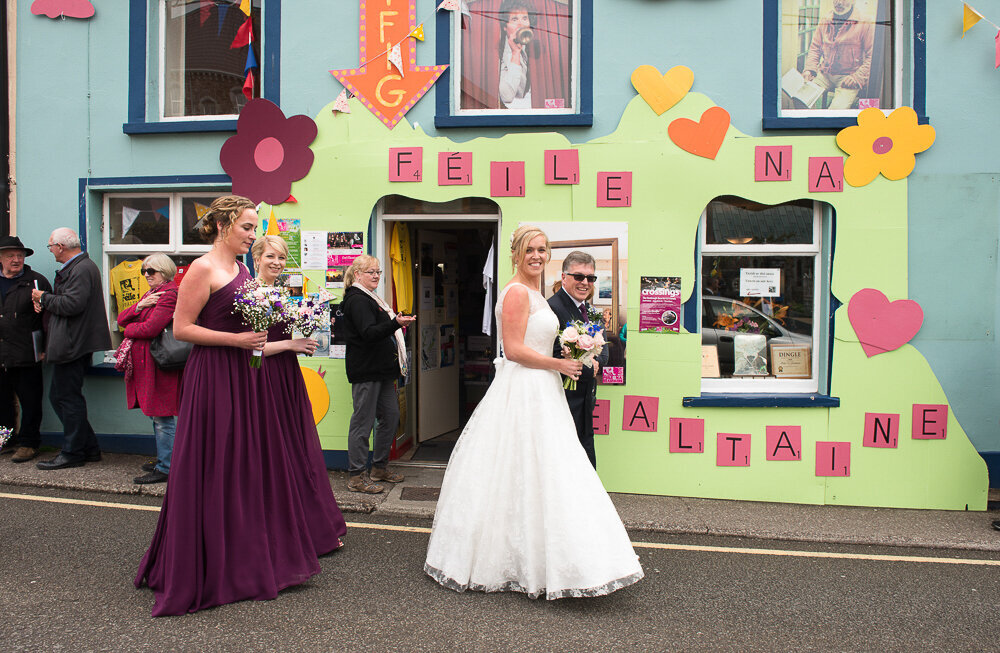 blonde bride wearing an a-line style wedding dress walking with her dad down Green street, Dingle followed by her bridesmaids wearing a-line, dark purple dresses