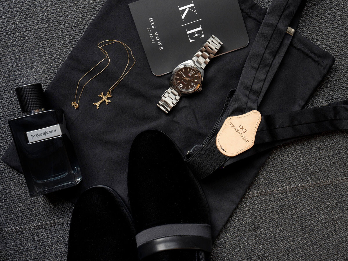 A flat lay of mens accessories, featuring a silver wristwatch, a gold cross necklace, a black cologne bottle, a pair of black velvet shoes, black suspenders, and a His Vows booklet on a dark fabric background.