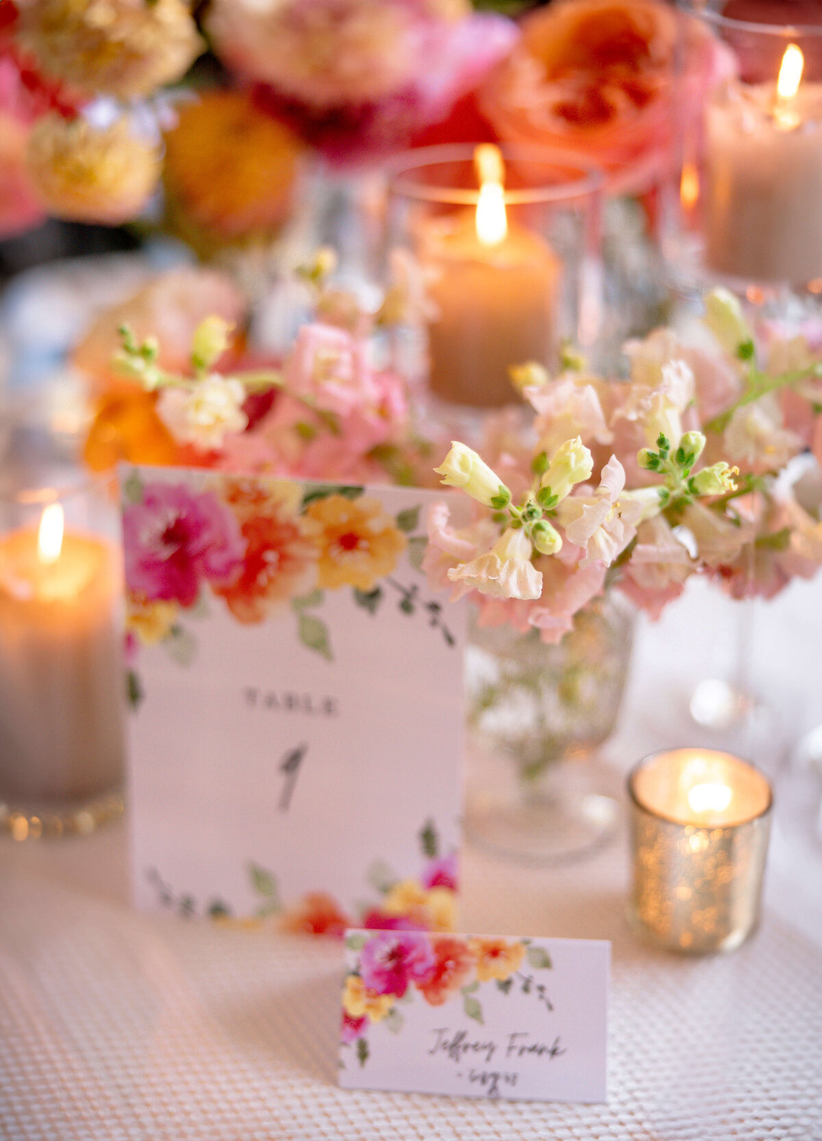 A beautifully arranged table setting featuring lit candles, floral arrangements, and a table number card with a floral design. Soft pink, orange, and yellow flowers add a touch of elegance and warmth to the decor.