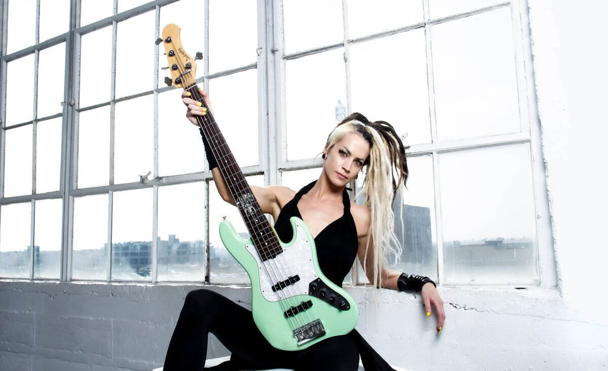 Female musician portrait Tanya O'Callaghan wearing black jumper sitting holding  pale green bass guitar against large window