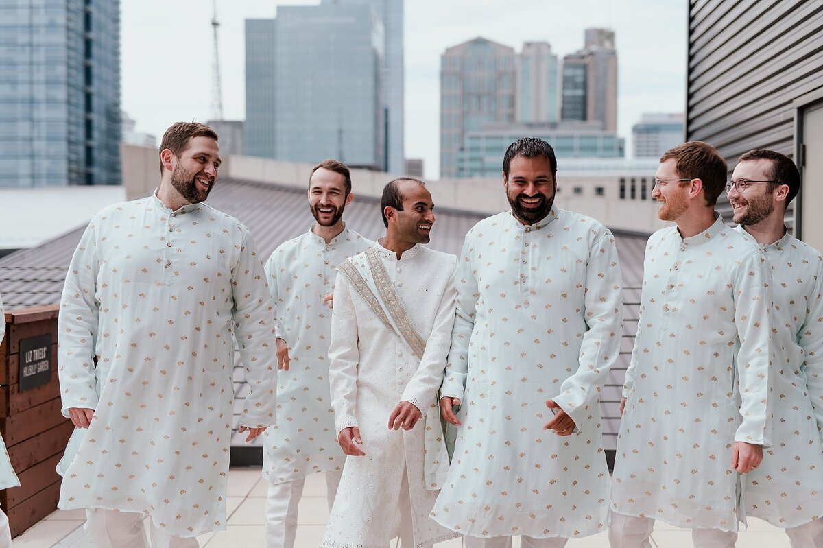 Indian Groomsmen wearing light blue and gold sherwani and they laugh with the Indian groom wearing a white sherwani with a light blue and gold dupatta with the Nashville Skyline in the background on the rooftop of the Country Music Hall of Fame.