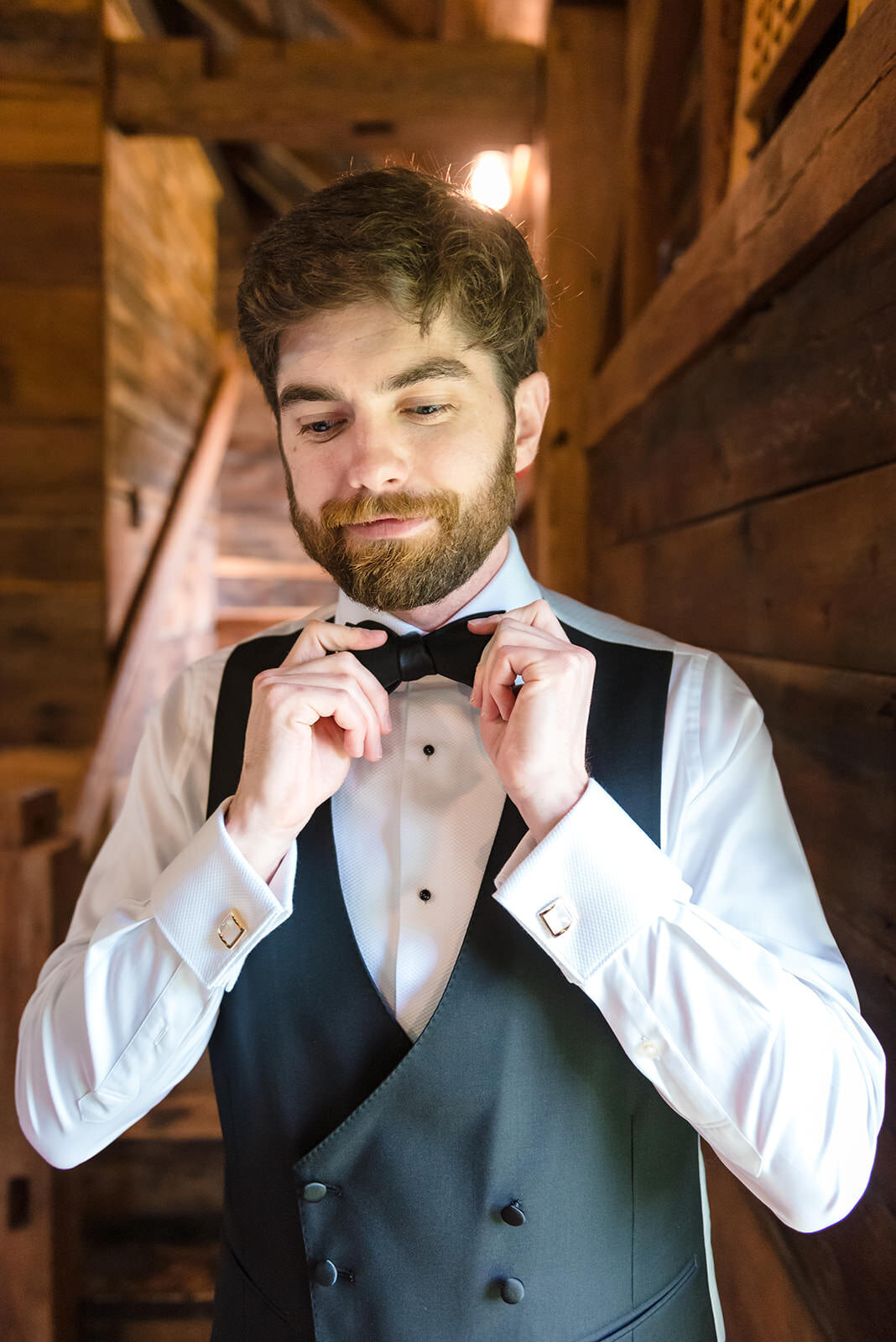 A man in a tuxedo adjusting his bow tie, with a rustic wooden backdrop.