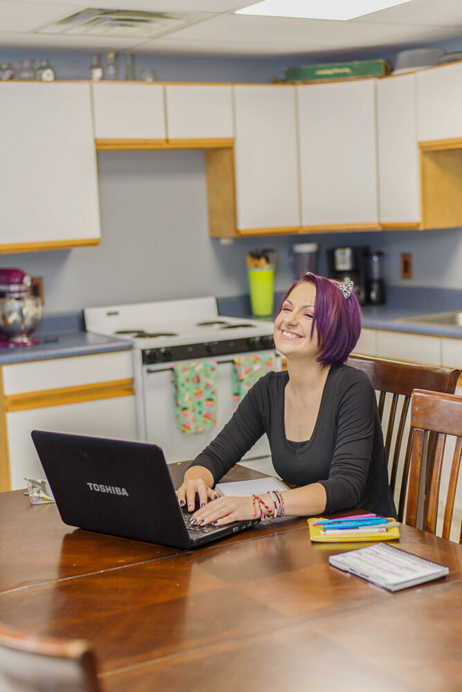 A woman with purple hair sits at a wooden table, smiling and looking away from her laptop in a room with kitchen cabinets.