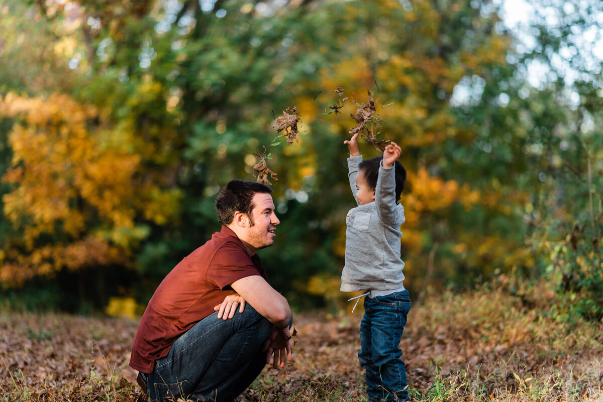 toddler boy throws leaves in the air while dad kneels to look at him