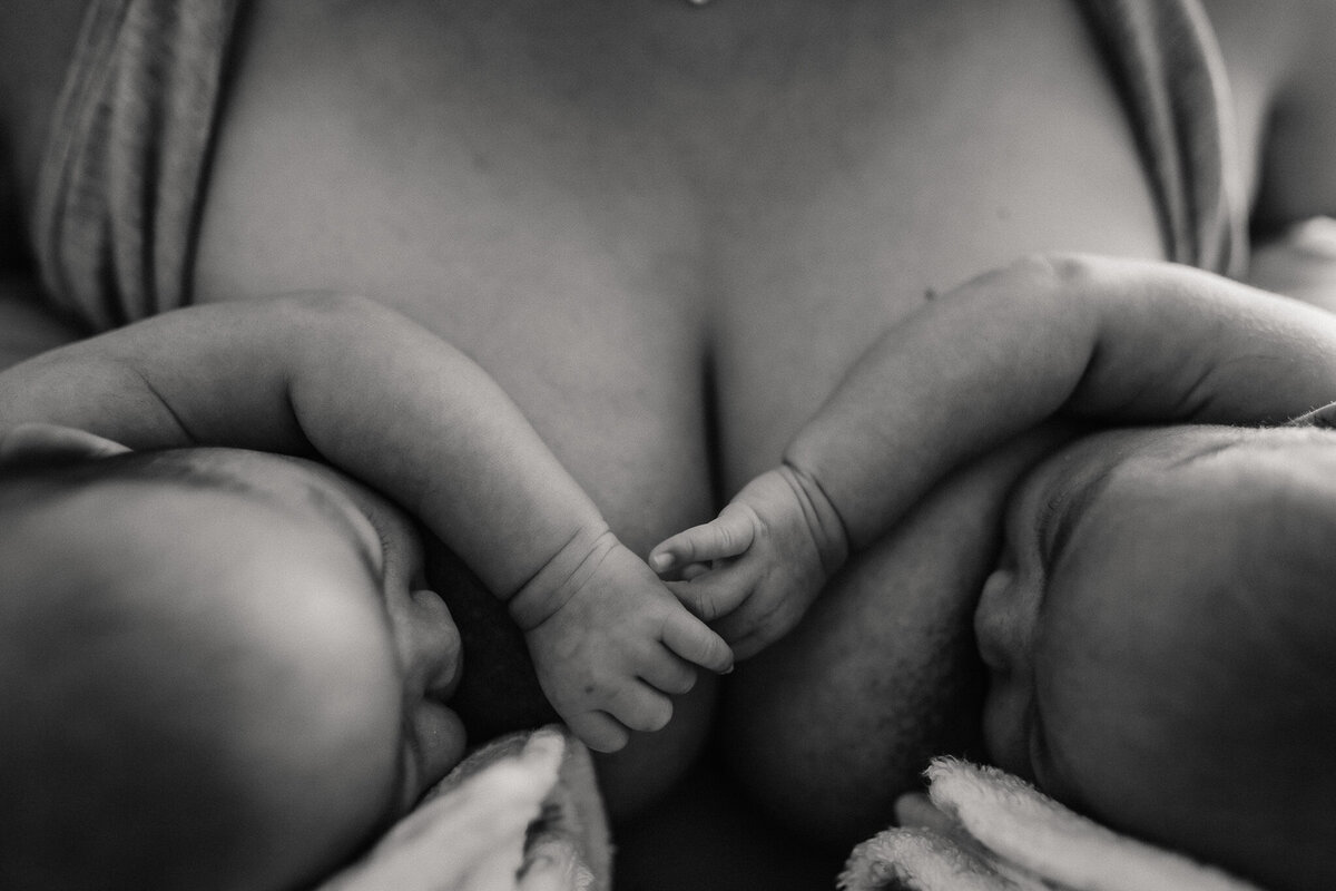 Lifestyle newborn photo of twins tandem nursing and holding hands. Image is in black and white.