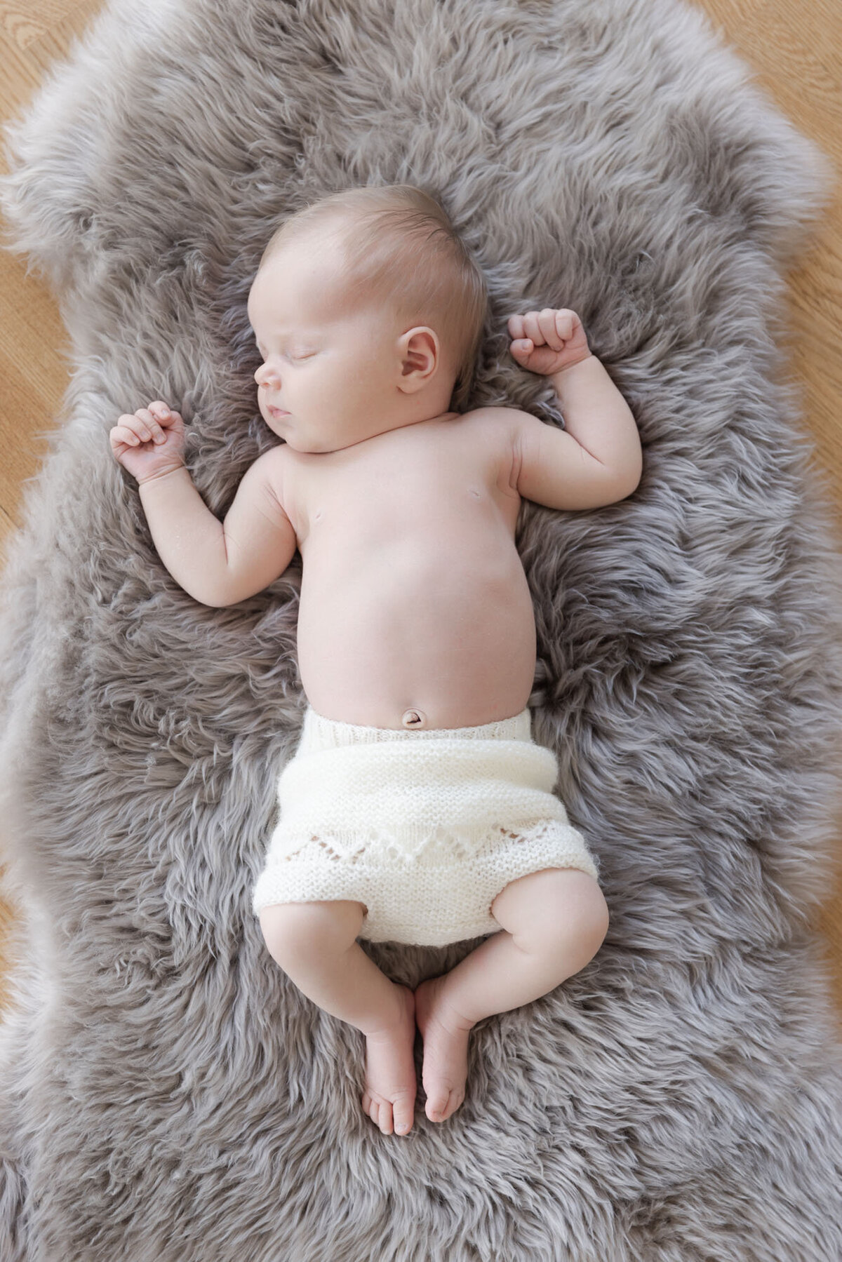 Livsstilsfoto tatt ovenfra og ned. Baby iført strikket bleieskjuler ligger på ryggen og sover på en grå ullfell.