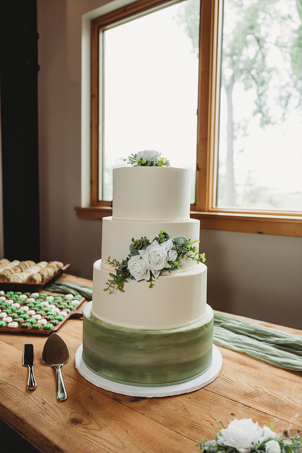 Mountain Sage Wedding cake at Camelot Ranch with sage green watercolor base, smooth buttercream icing, and white flower accents on a rustic wood table.