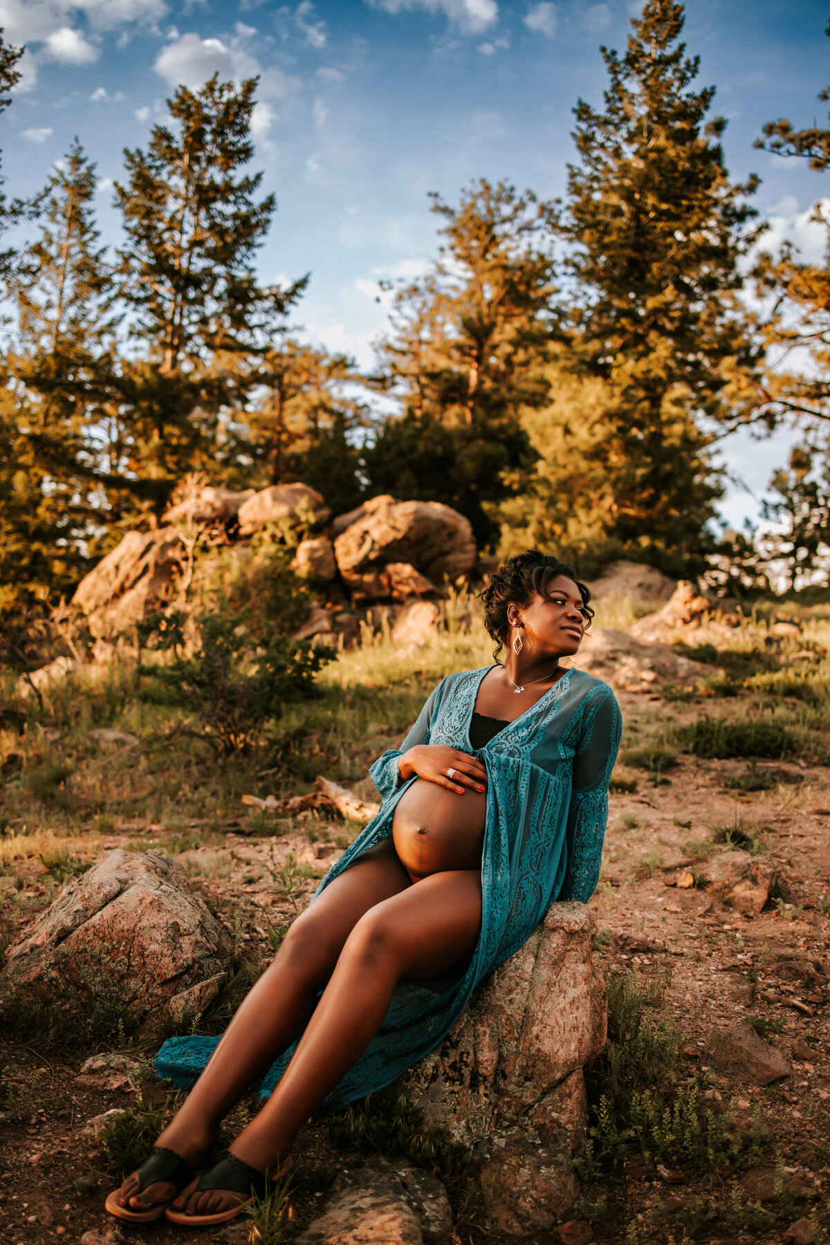 african american pregnant mom sitting on a rock in the mountain s with her bare belly showing through blue dress