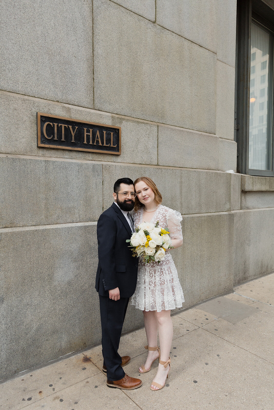 couple just married standing under Chicago city hall sign. Looking at camera and smiling, bride is holding a yellow bouquet.