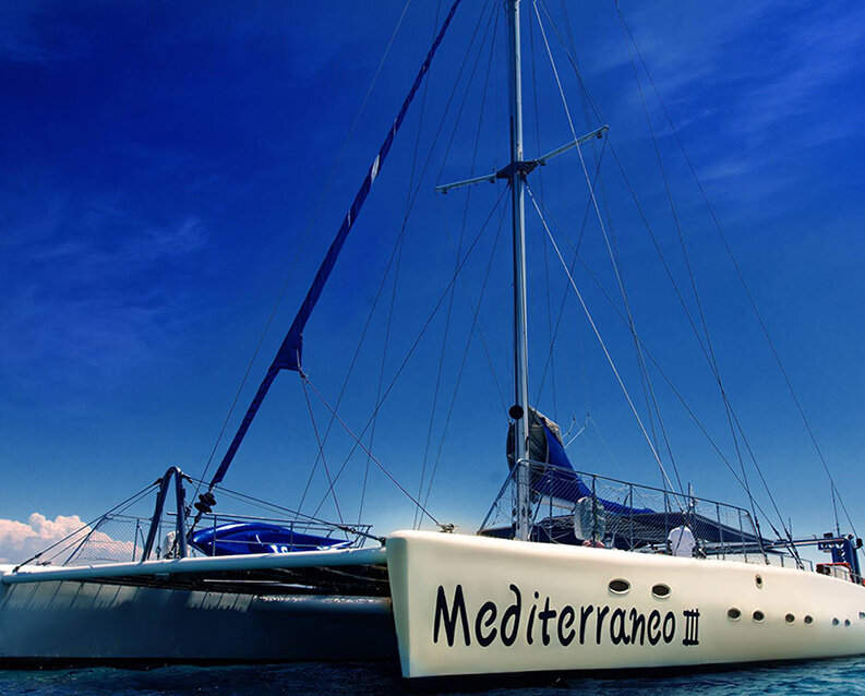 A white catamaran sails along the med against a pure blue sky
