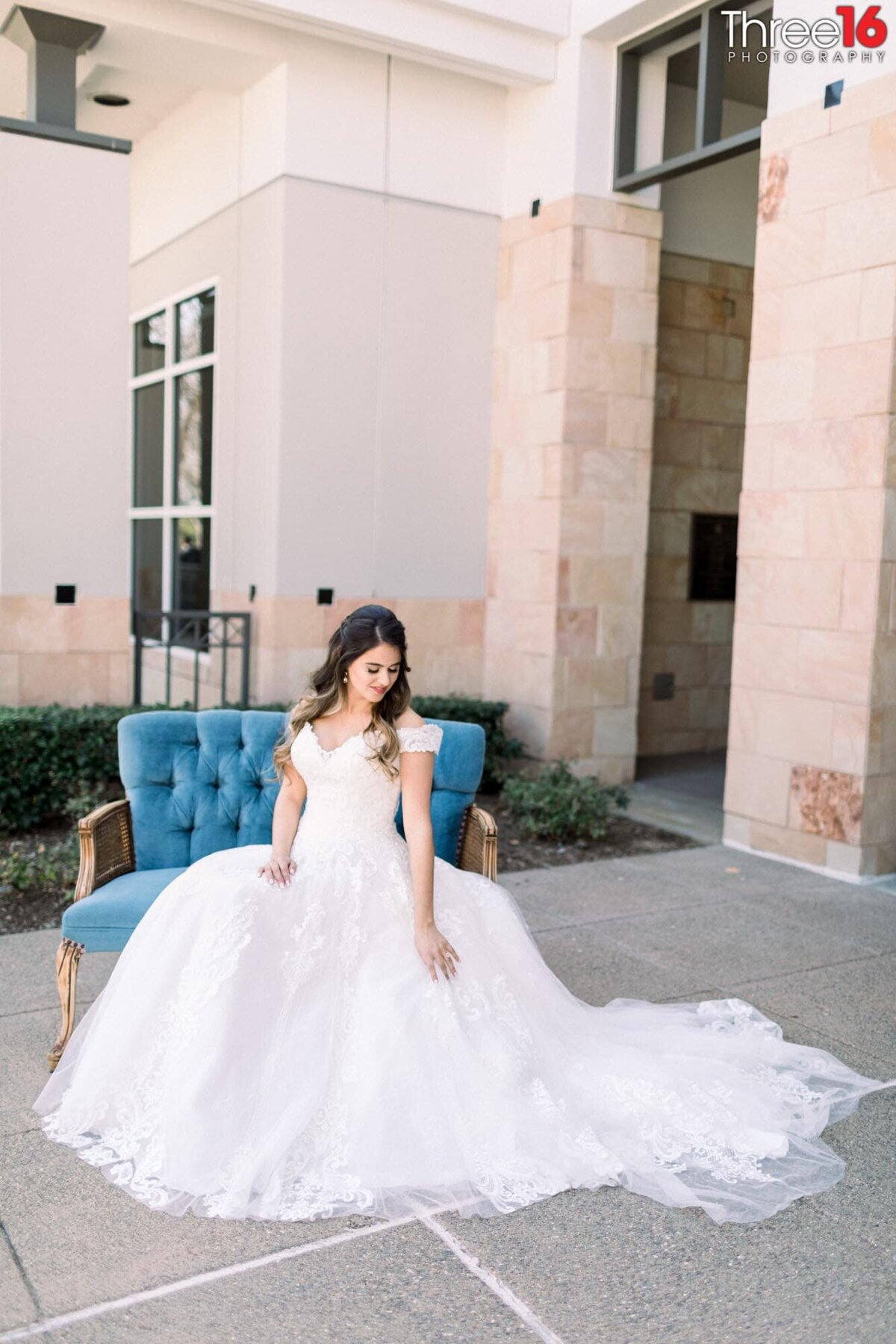Bride poses before the ceremony sitting on a blue love seat with her dress fanned out in front of the entrance at the Yorba Linda Community Center