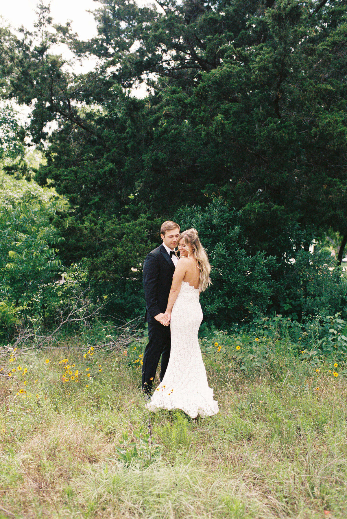 A couple embraces in a grassy field, surrounded by wildflowers and lush greenery. The woman wears a white dress; the man, a dark suit. The scene is serene and romantic.