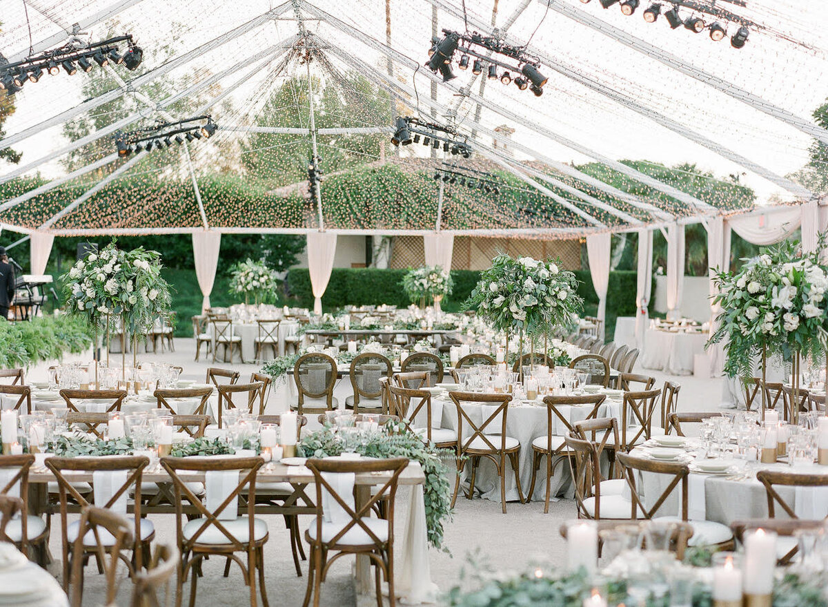 clear tented wedding reception space with round tables, tall centrepieces filled with green florals and plants, round tables, and dark wooden chairs.