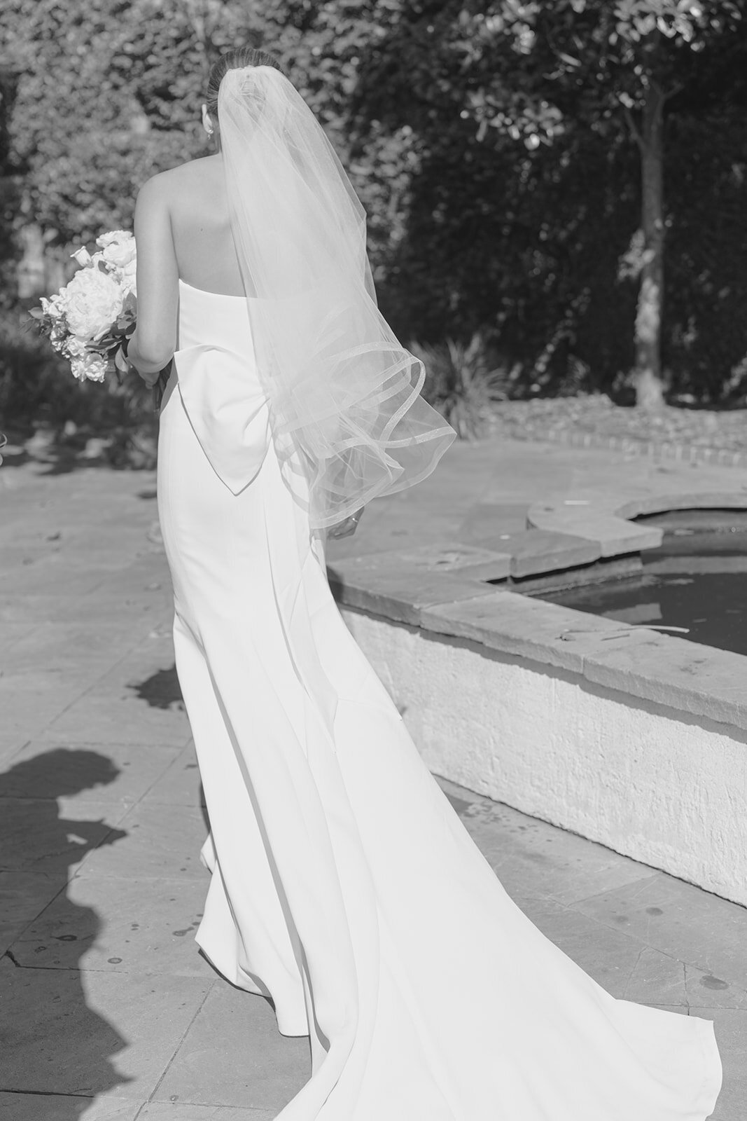 Wedding veil blows in the wind as bride walks to outdoor wedding ceremony at William Aiken House.  Black and white destination wedding photographer. Kailee DiMeglio Photography.
