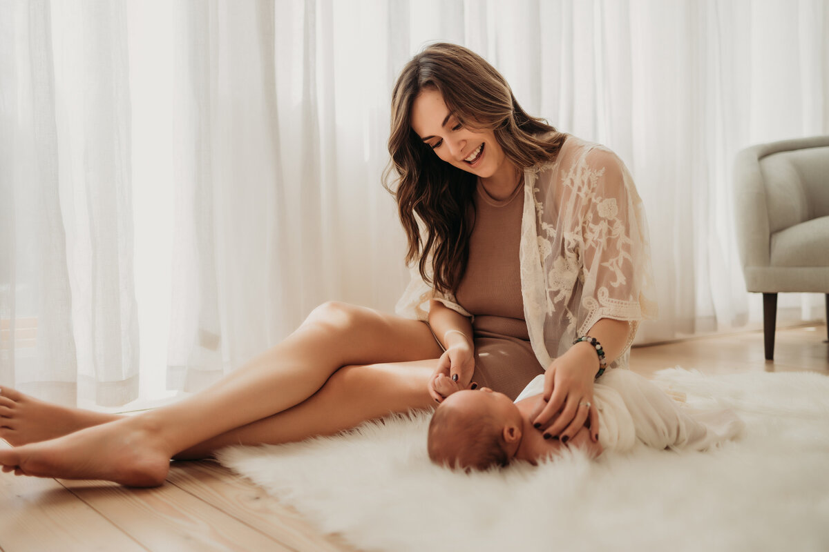 new mom looking down at baby sleeping on a white rug and holding her hands