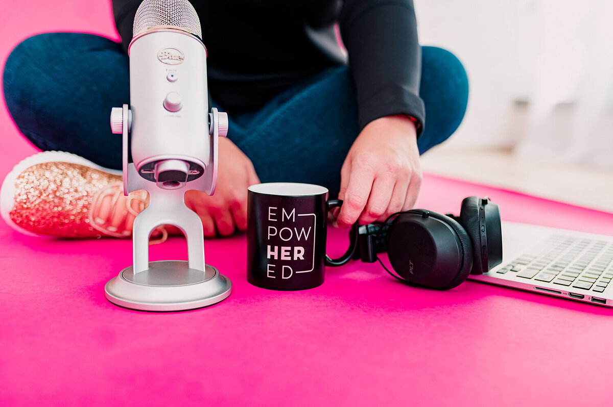 woman sitting on hot pink floor with a podcast mic, black mug, headphones and laptop