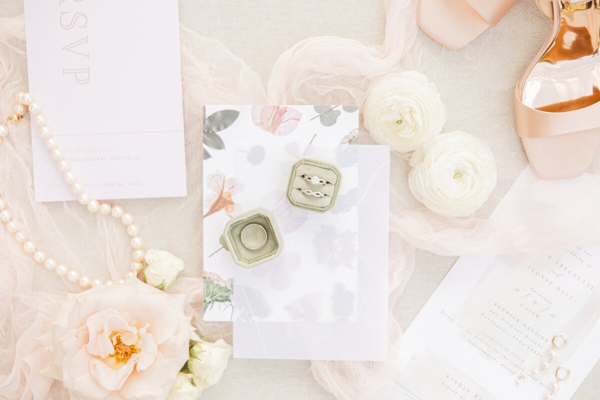 Flat lay of wedding accessories including invitations, RSVP cards, a pair of shoes, pearls, white flowers, and a ring box arranged elegantly on a soft fabric background by a wedding planner in Des Moines