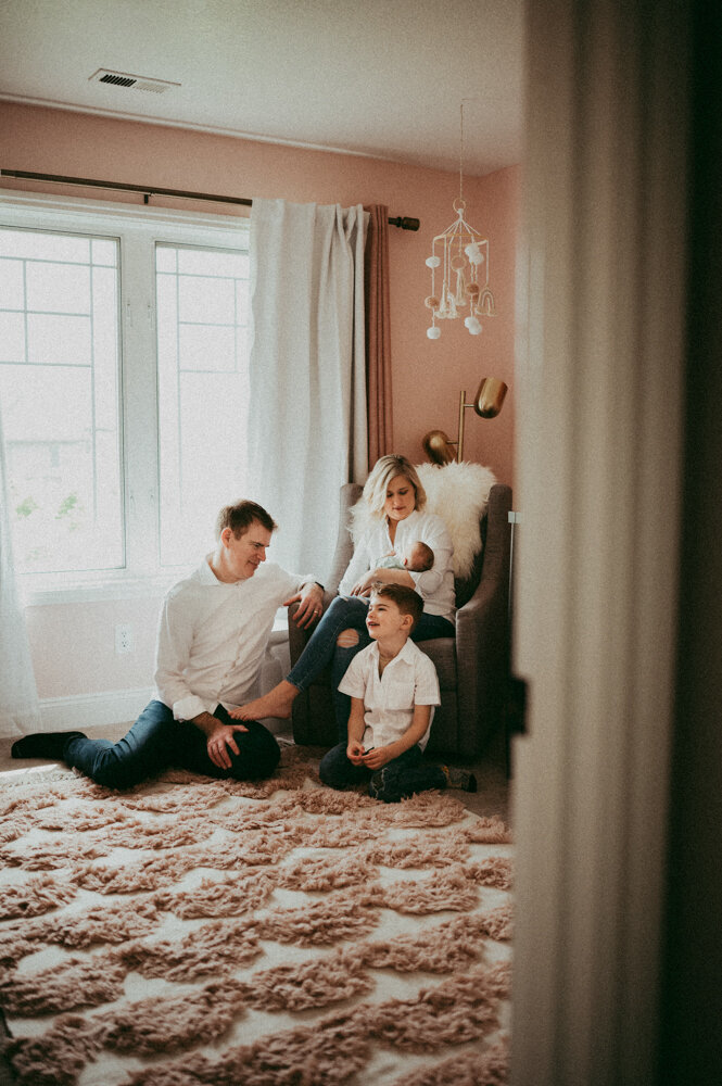 Cozy newborn lifestyle session in a softly lit nursery—mother nursing baby in a rocking chair while father and older sibling sit close, sharing a calm family moment.