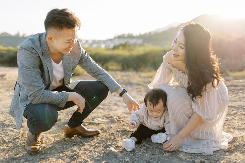 Family of three in a grassy outdoor photoshoot with baby playing with sand.