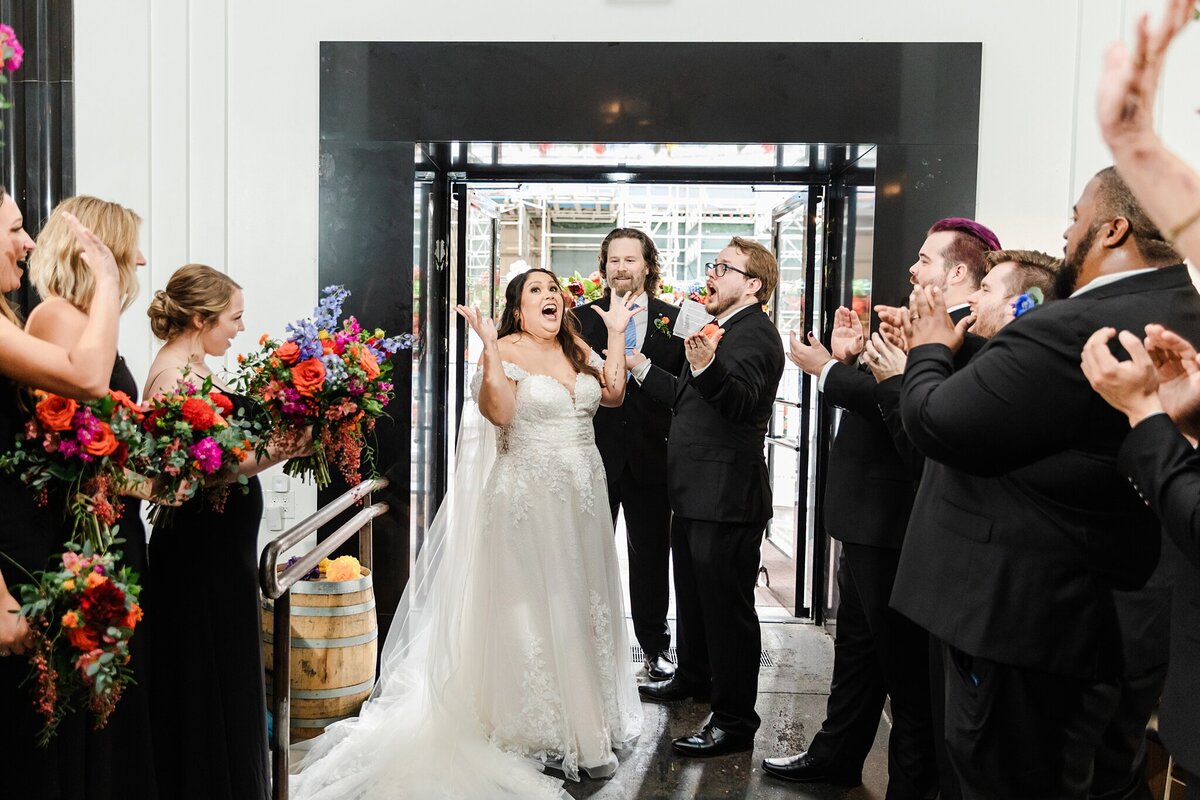 A bride and groom acting joyfully shocked at the conclusion of their wedding ceremony at the Pegasus City Brewery in Dallas, Texas. Both of them are holding up their hands in pretend shock (but genuine joy). The bride is on the left and is wearing a long, flowing, white dress and long veil. The groom is on the right and is wearing a dark suit with a boutonniere. Their officiant stands behind them while their wedding party celebrates on either side.
