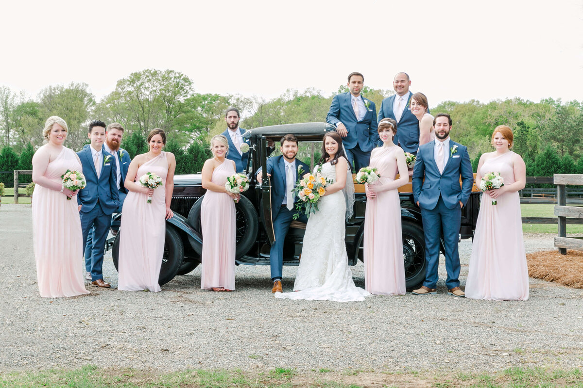 A bridal party in pinks and blues pose in front of a vintage car.