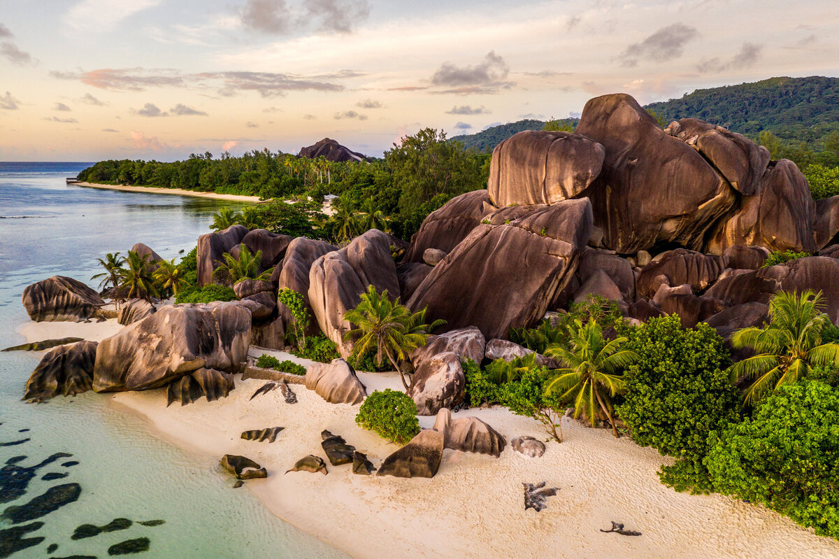 Aerial view of a tropical beach featuring large, smooth boulders nestled among lush green palm trees. The shoreline curves gently with clear turquoise water. The sky is tinged with pastel hues of sunset, and a forested hill rises in the background.