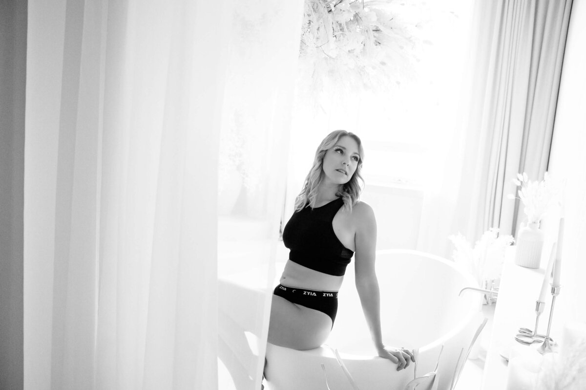 Black and white photo of a Blonde woman wearing a black Zyia bra and underwear in a bathtub for her Burlington Boudoir Photography Session