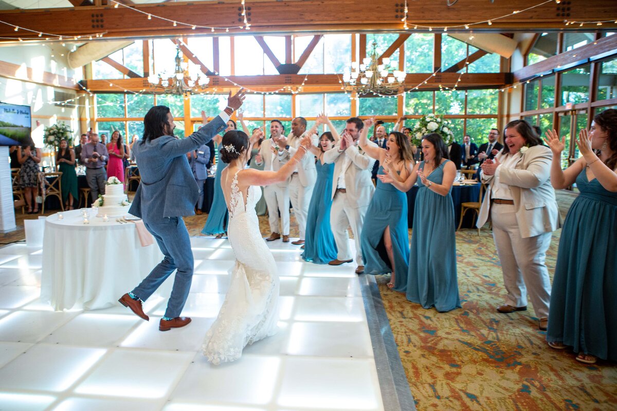 Bride and groom dancing joyfully with guests clapping around them in a bright, elegant hall decorated with chandeliers at the Park Farm Winery weddings.