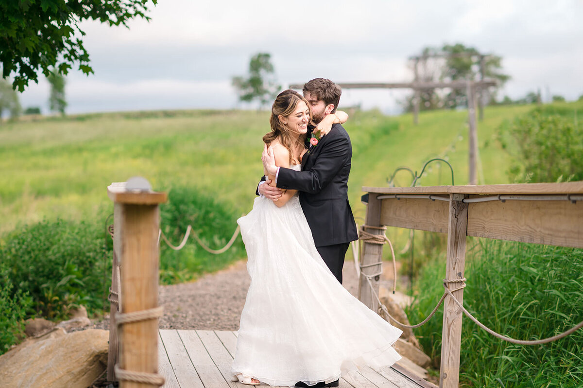 A bride and groom share a tender moment on a bridge in a pastoral setting, with a wooden structure in the distance.