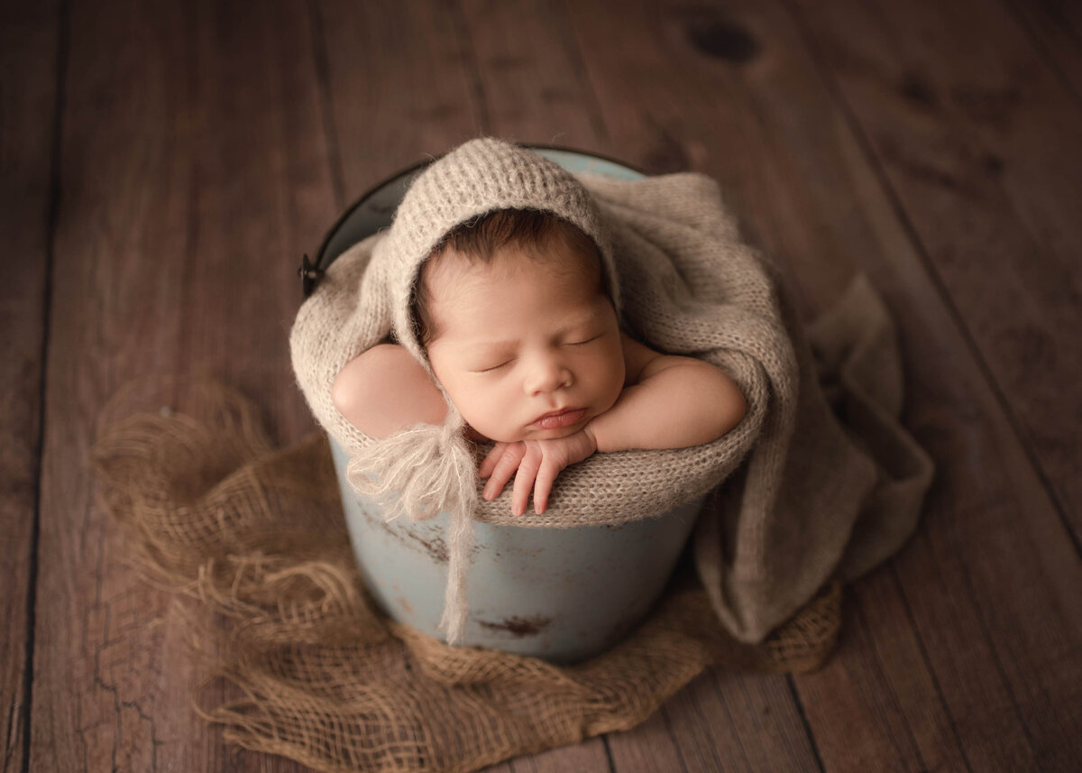 newborn Baby boy in bucket with knit wrap and bonnet on hard wood floor