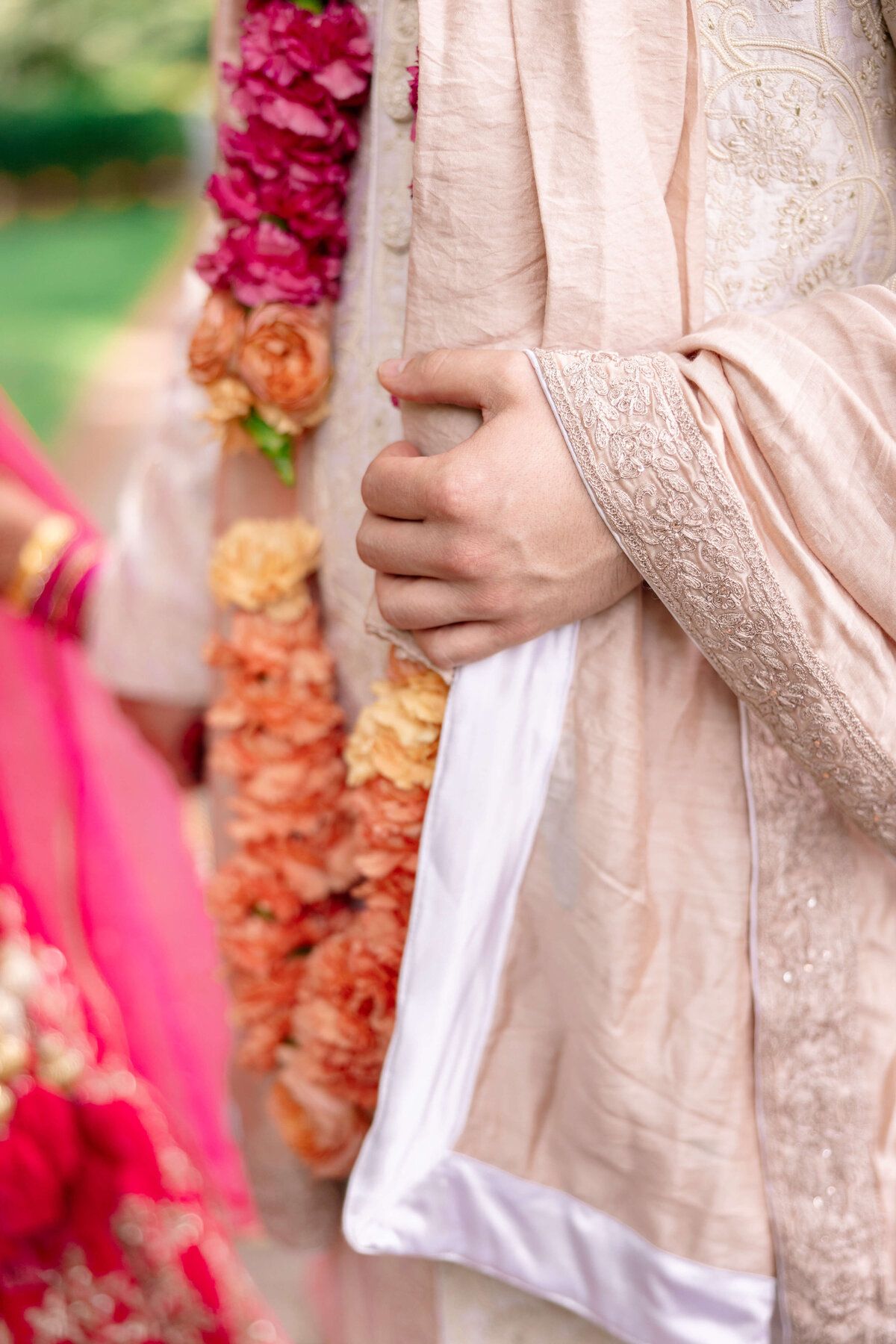 Close-up of a person in a traditional beige outfit with intricate embroidery, holding a garment. They are wearing a garland of orange and pink flowers. Another person in pink attire stands nearby, and the background is a blurred outdoor setting.