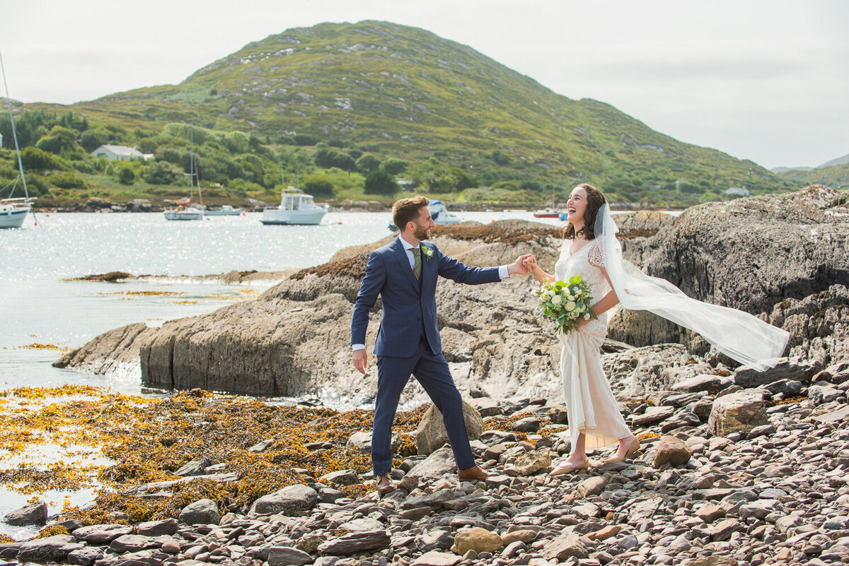 Bride wearing sequenced, vintage wedding dress and veil standing on pebbled beach laughing with groom wearing a navy suit and green, tweed tie