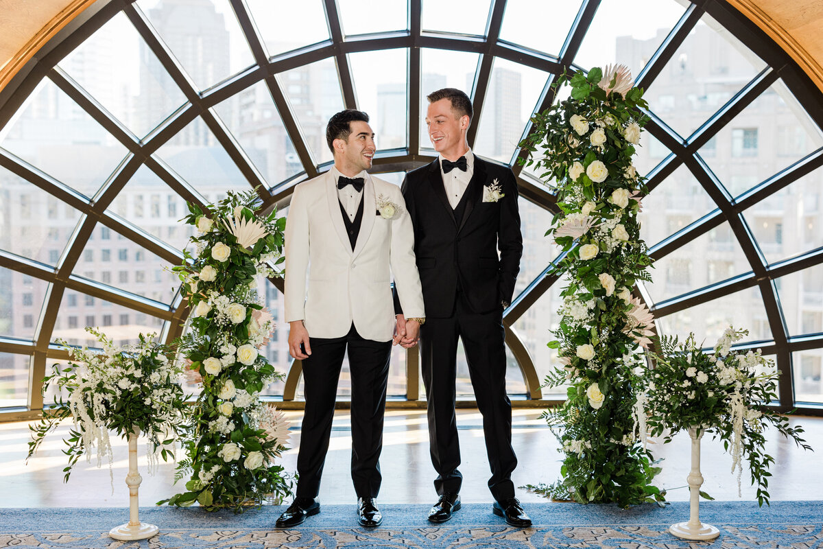 A portrait of two grooms posing at the alter of a wedding ceremony at the Crescent Club in Dallas, Texas. The groom on the left is wearing a tuxedo with an all black tuxedo except for the white jacket and boutonniere. The groom on the right is wearing an all black tuxedo and boutonniere. They are both holding hands  while smiling at each other and are surrounded on either side by large floral arrangements.