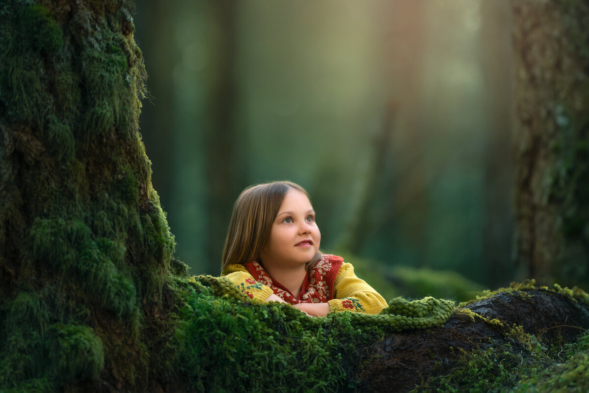 A young girl pops up from behind a moss-covered log during her stylized, fantasy portrait session.