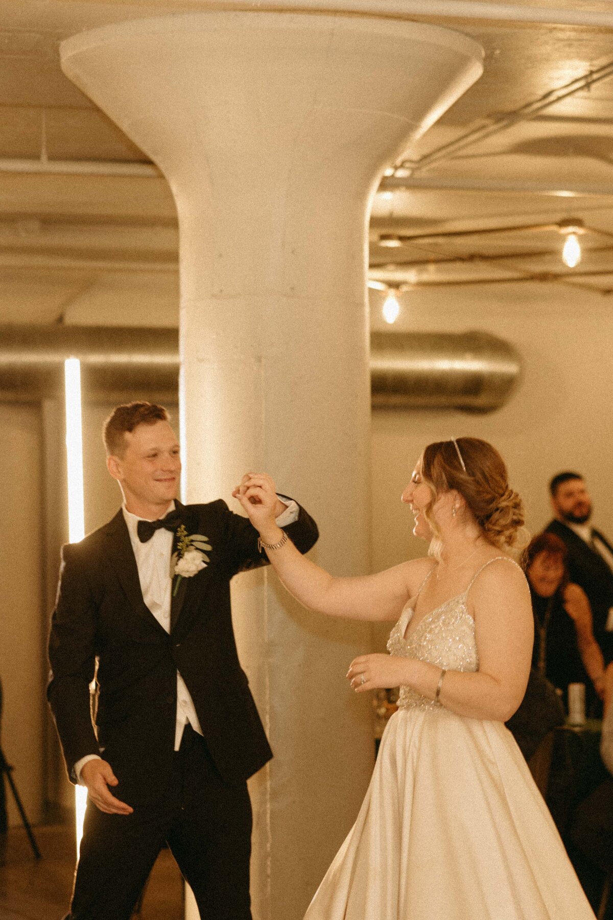 A bride and groom dancing joyfully in a warmly lit hall at a Park Farm Winery wedding, with guests watching in the background. The groom is adjusting the bride's sleeve, both smiling.