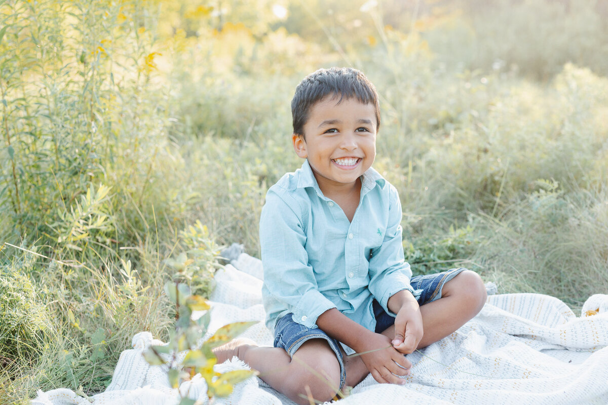 Little boy sits in a field on a white blanket wearing a button up blue shirt.