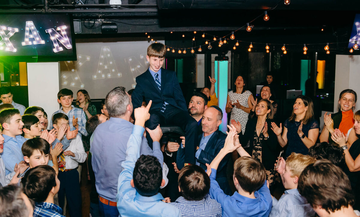 A boy smiles big while lifted on the dance floor in a chair