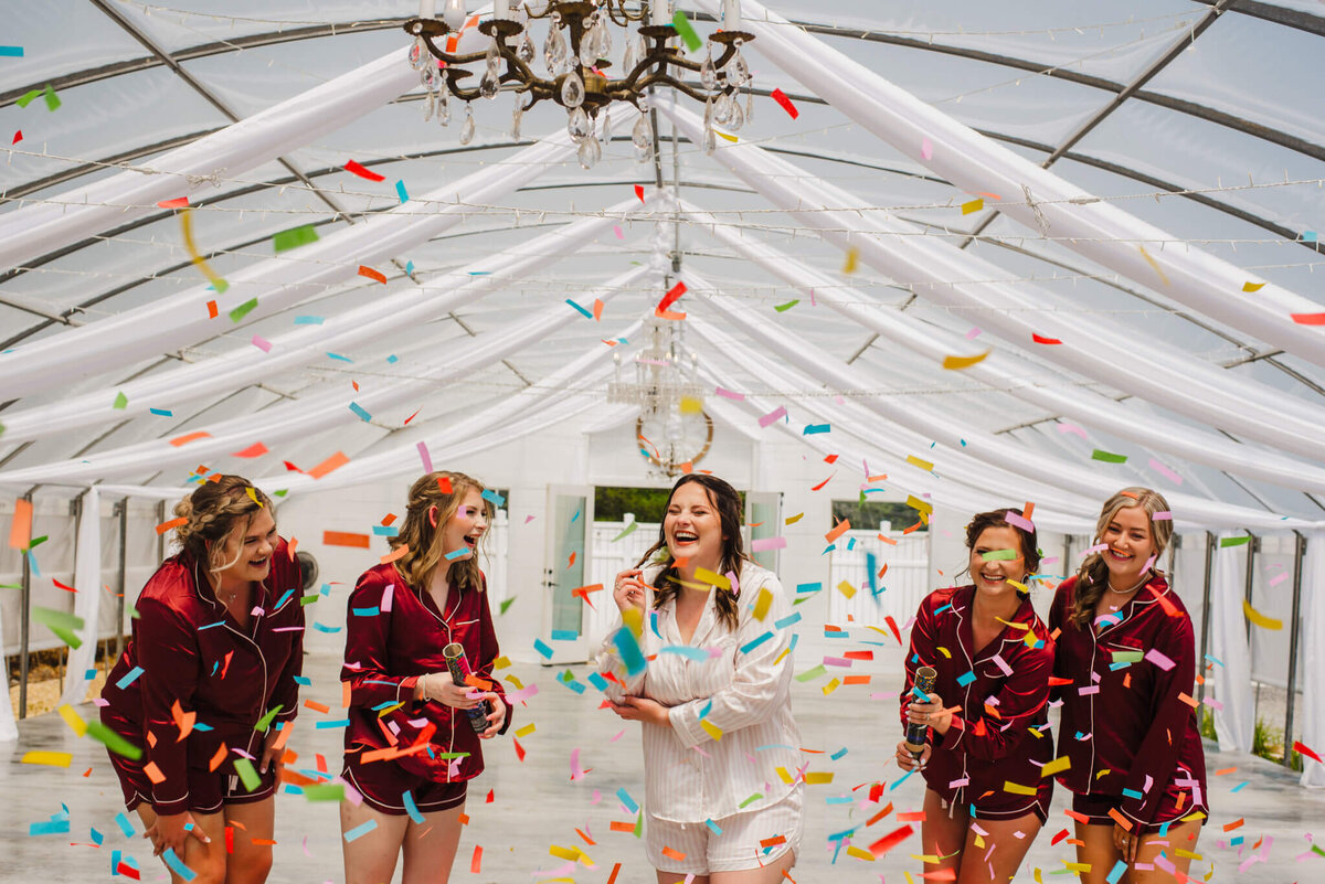 Photo of a bride in white pajamas and bridesmaids and maroon pajamas popping confetti inside of a white greenhouse