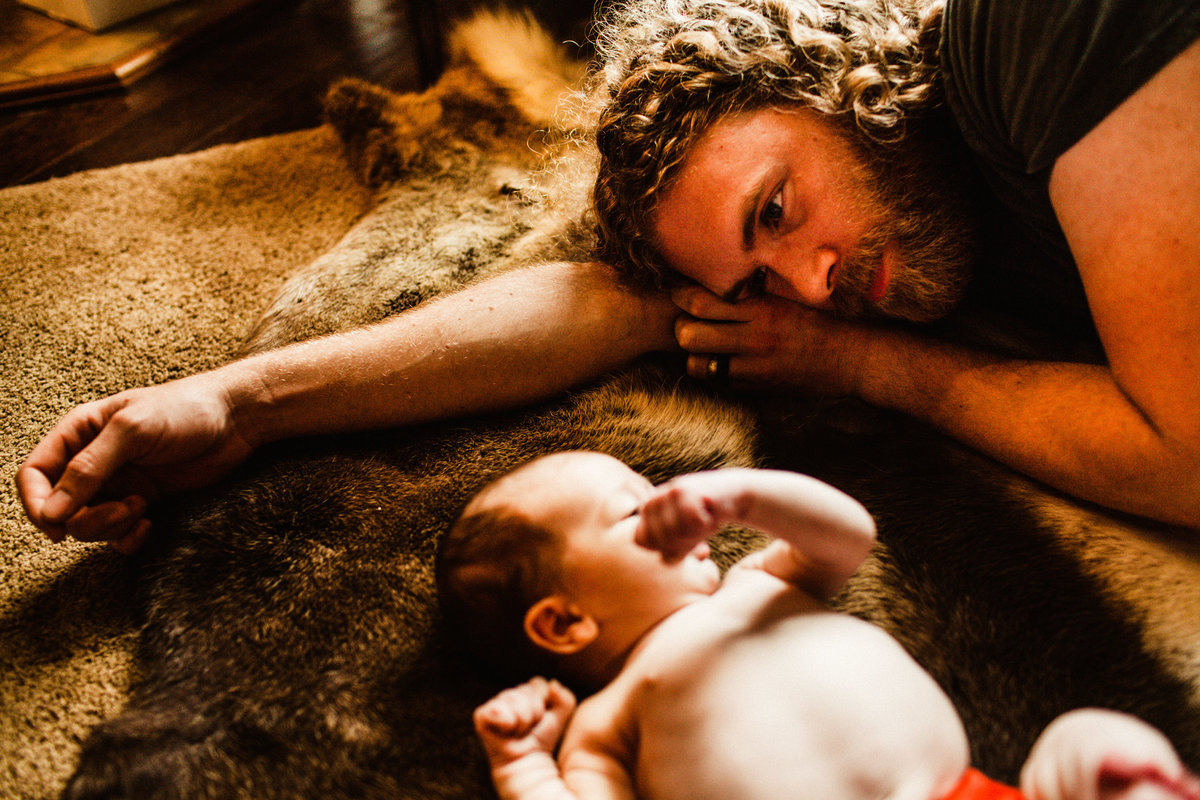 father lays on fur rug looking at newborn son