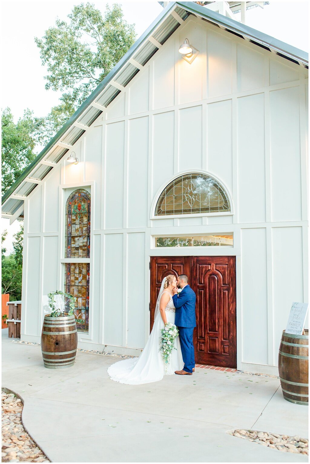 bride and groom portrait kissing outside white chapel with wooden doors and whiskey barrel flowers